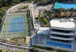 A blue tennis court alongside a modern building with a swimming pool.