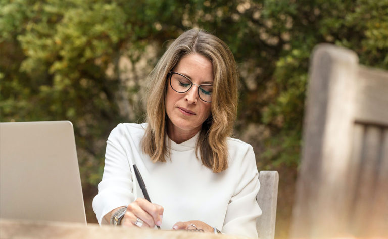 A woman in a white dress in the garden, writing notes in front of its laptop.