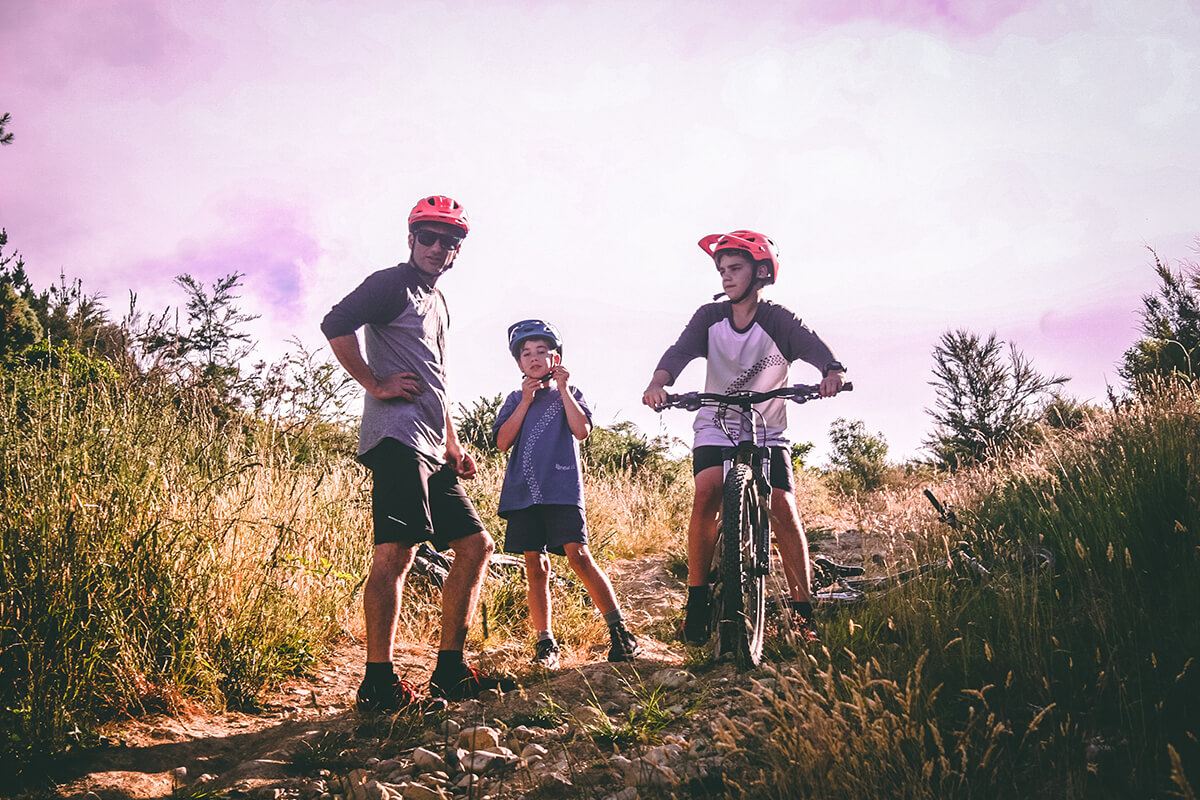 A father with his two sons riding a bicycle wearing orange helmets.