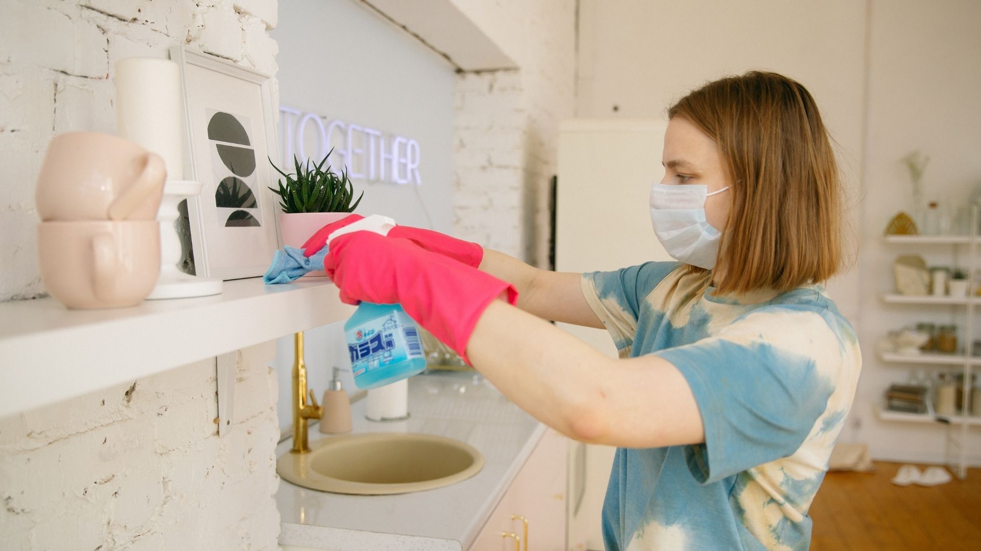A woman with a blue t-shirt, face mask, and pink gloves, cleaning the surface in the kitchen.