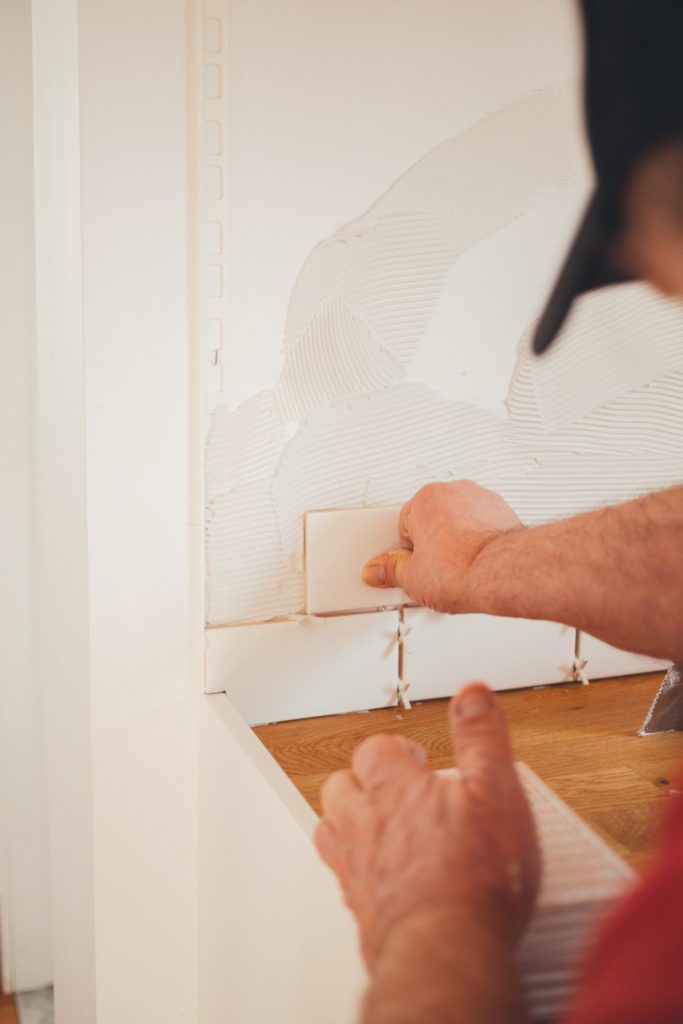 A worker with a black hat lays white tiles in the kitchen.